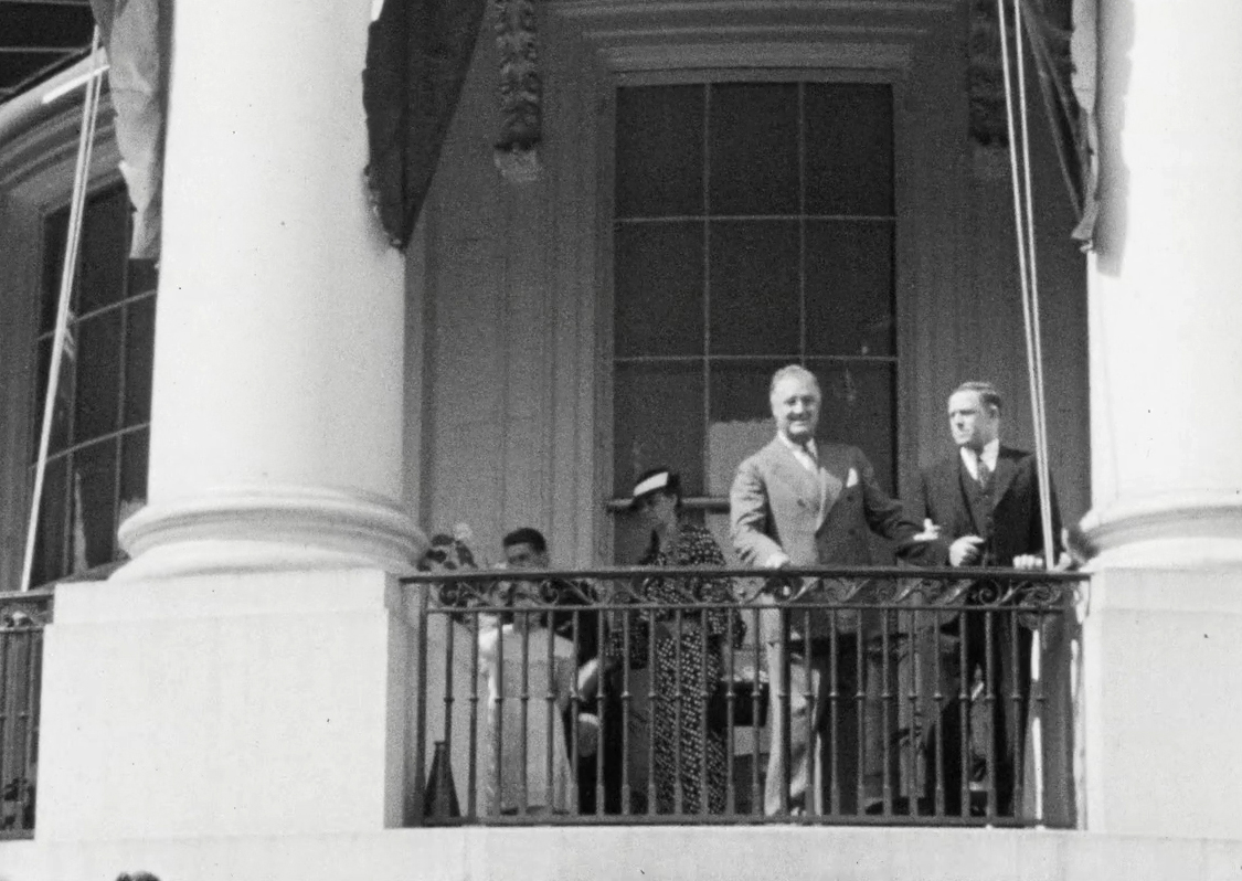 FDR stands at at the railing of the South Portico of the White House, bodyguard Gus Gennerich at his arm. 